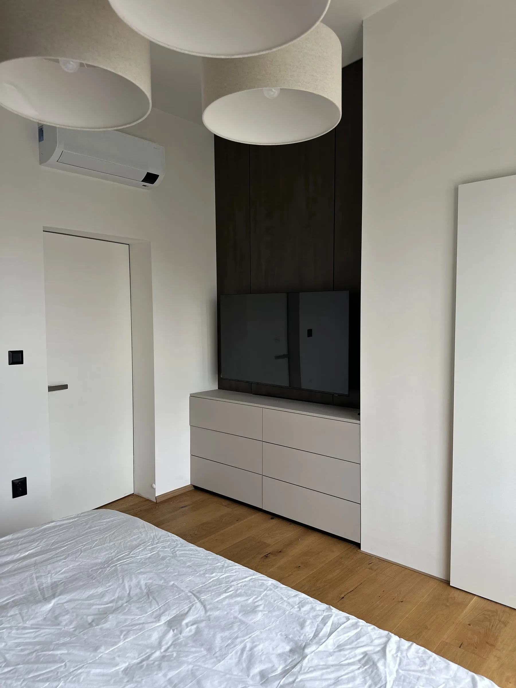 Spálňa: tmavé panely za televízorom, biela komoda a dve svietidlá nad posteľou